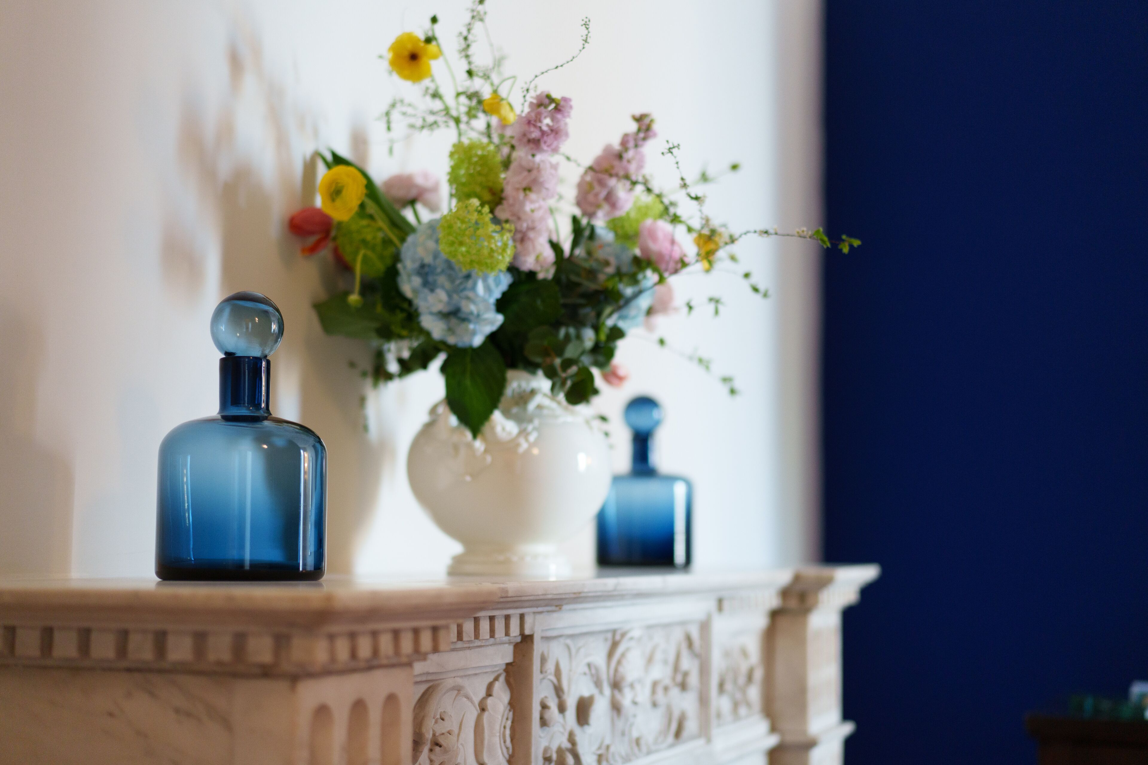 Ein Marmor-Kamin mit einer weißen Vase, die eine Blumenkomposition und zwei blaue Flaschen enthält.