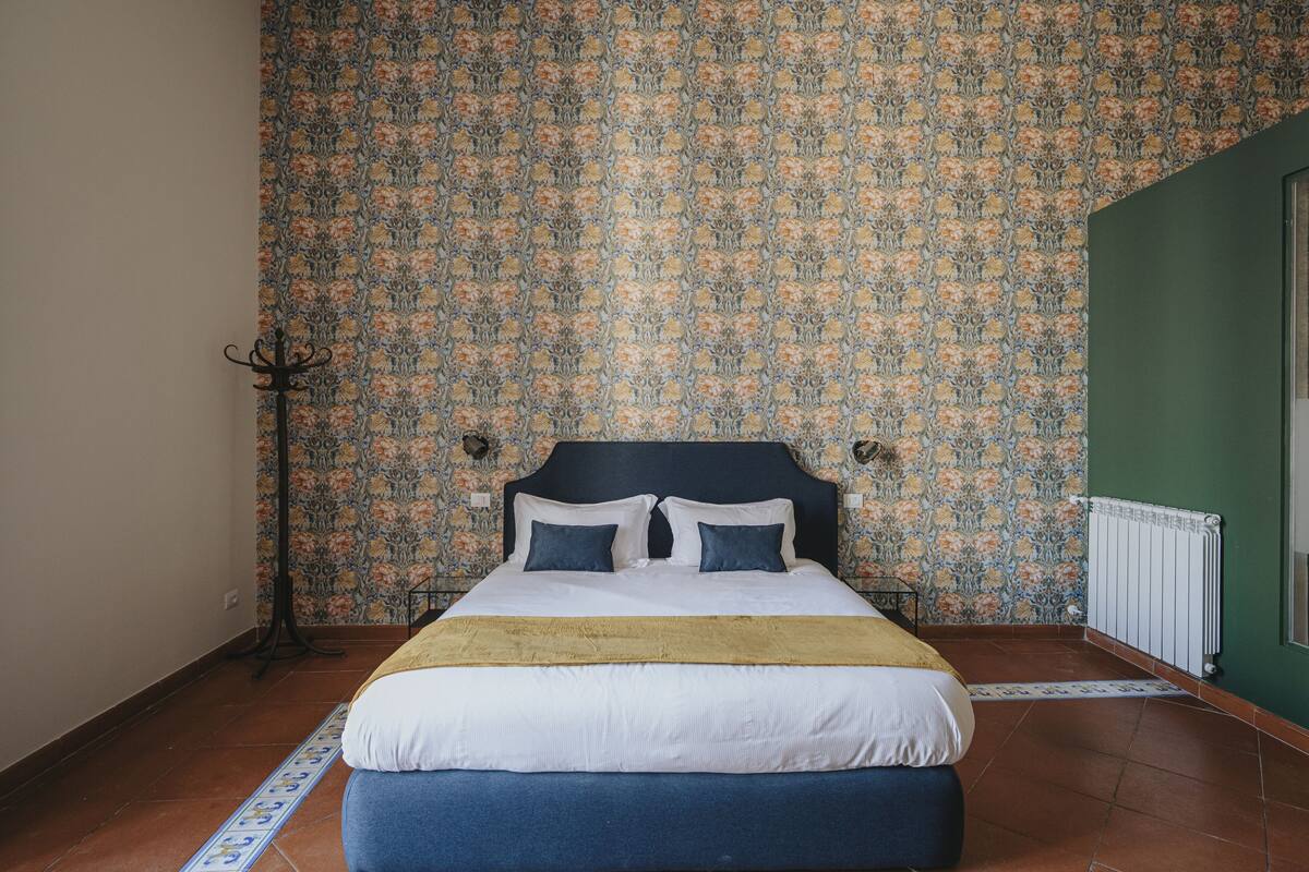Ein Schlafzimmer mit gemustertem Wandpapier, einem Bett mit weißer und blauer Bettwäsche, einem Heizkörper und einer grünen Wand.