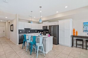 A modern kitchen with white cabinets, a central island, blue chairs, and a dining area. - Fun 2BR Retreat! Theme Room, Lake View near Disney (Kissimmee)