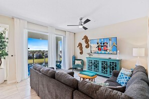 A living room with a grey sofa, a teal cabinet, and a television displaying animated characters. - Fun 2BR Retreat! Theme Room, Lake View near Disney (Kissimmee)