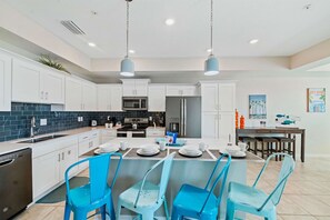 A modern kitchen with white cabinets, a central island with seating, and a dining area with blue chairs. - Fun 2BR Retreat! Theme Room, Lake View near Disney (Kissimmee)
