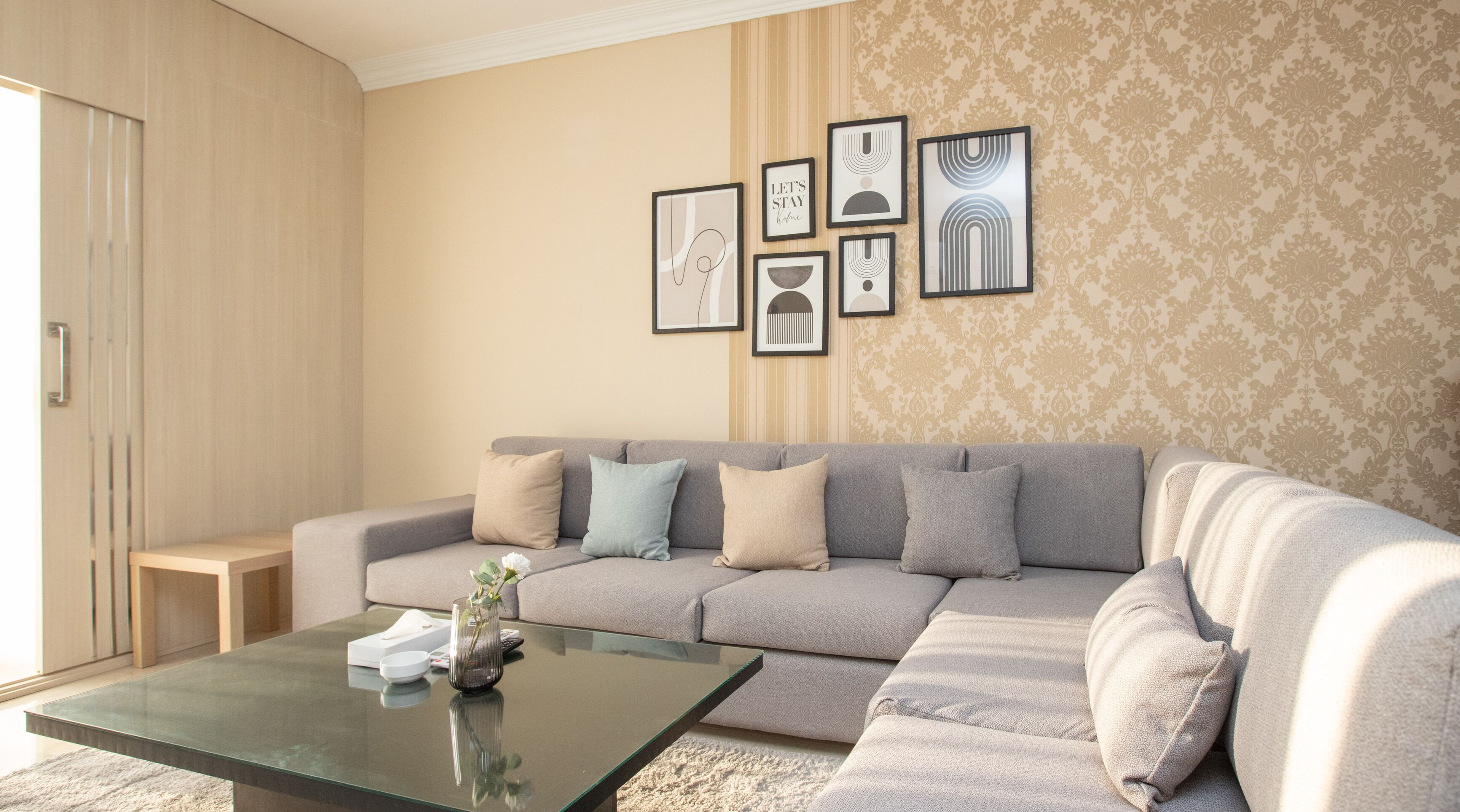 A living room with a grey sectional sofa, a glass coffee table, and a patterned wallpaper.