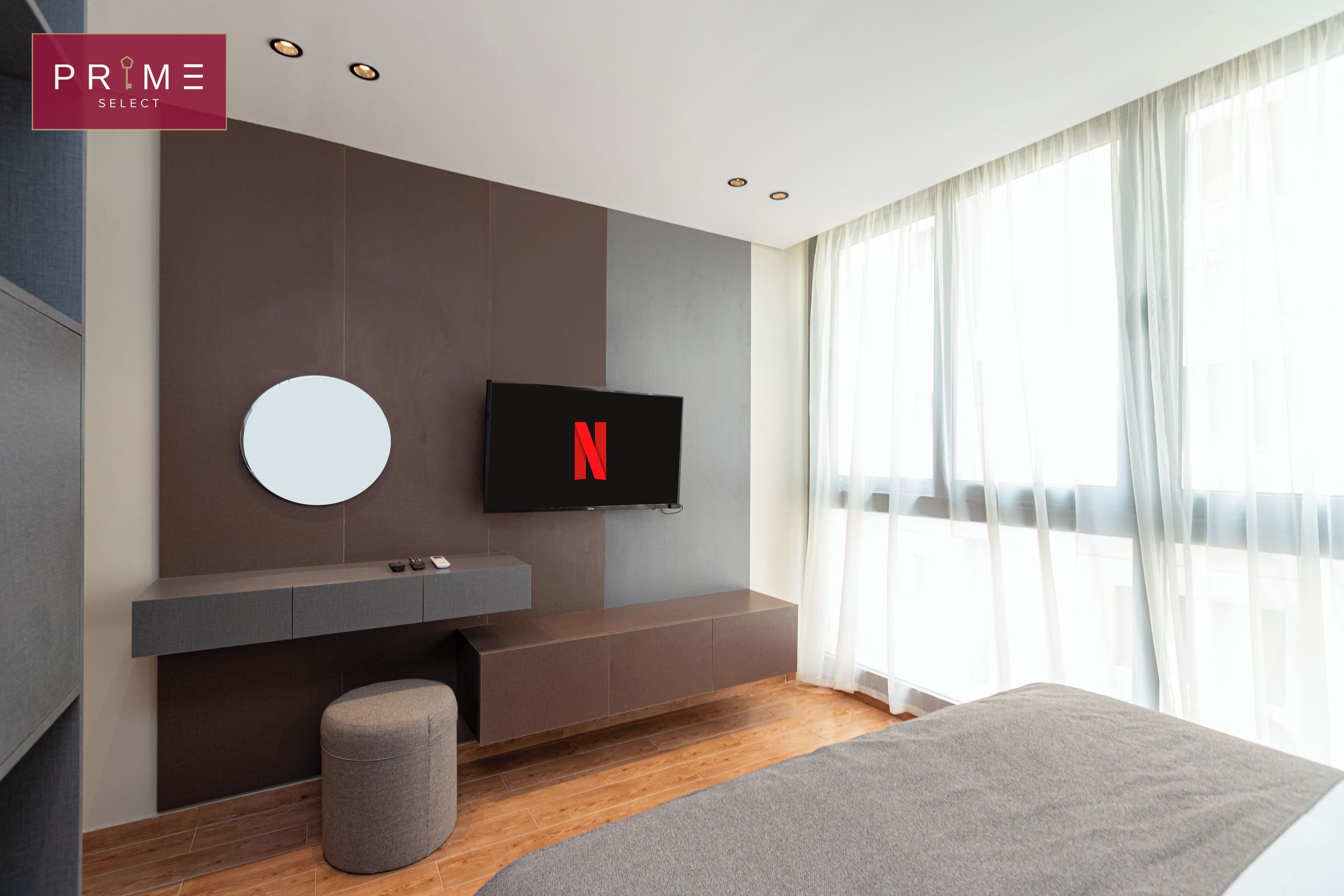 Ein modernes Schlafzimmer mit einem großen Bett, einem an der Wand befestigten Fernseher, einem runden Spiegel und einer Sitzbank mit Stauraum.
