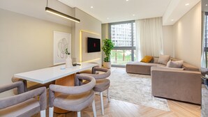 Un salon moderne avec une table à manger blanche, des chaises grises à revêtement en tissu, un canapé beige et une grande fenêtre donnant sur un paysage urbain.
