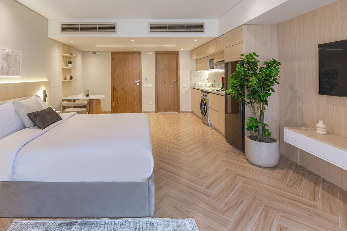 A modern hotel room with a large bed, a small desk, a washing machine, and a potted plant.