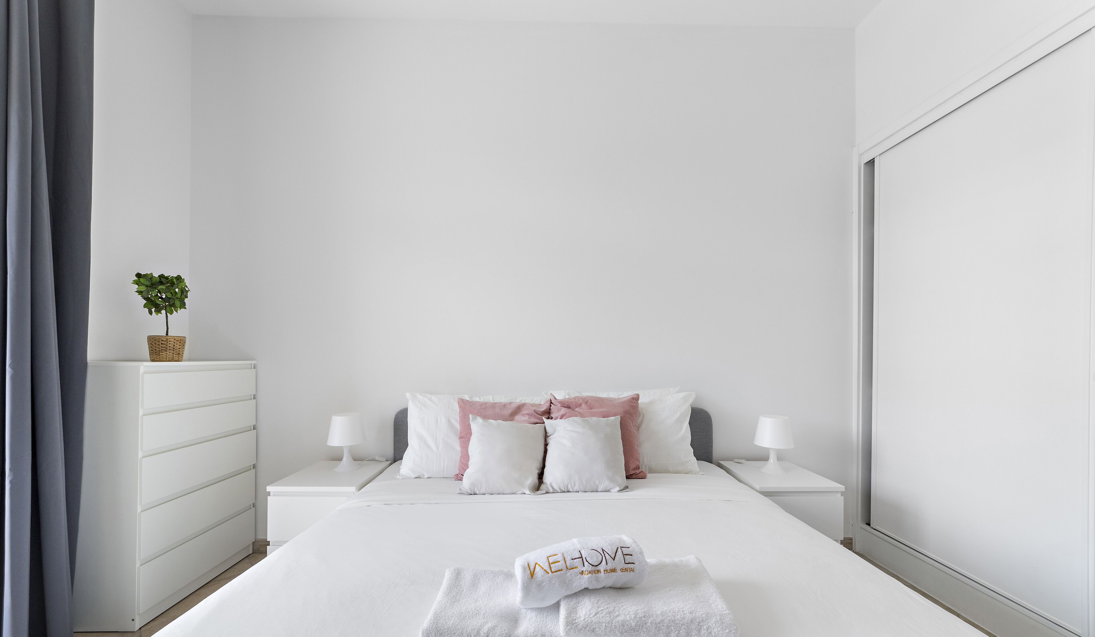 A minimalist bedroom with a bed, white nightstands, a bedside table, a small potted plant, and a towel with "WELCOME" on it.