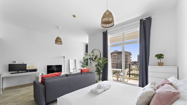 Une chambre d’hôtel moderne dotée d’un grand lit, d’un canapé gris, d’une télévision à écran plat et d’un balcon offrant une vue sur la ville.