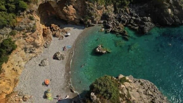 Ein felsiger Strand mit klarem türkisblauem Wasser und einigen Sonnenschirmen.