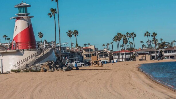 A beach with a red and white lighthouse, palm trees, and beach houses. - 114 31st A | Rare 1 bdr On Peninsula 3 Houses To Sand (Newport Beach)