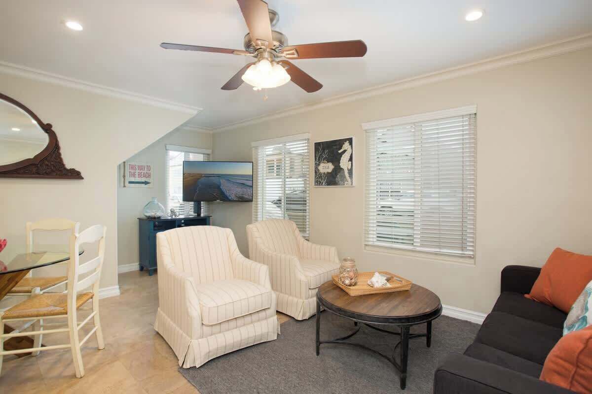 A living room with a ceiling fan, a dining table with chairs, a sofa, and a coffee table. There are two armchairs, a TV, and a window with blinds.