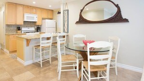 A compact kitchen with wooden cabinets, a microwave, and a refrigerator. There is a dining area with a glass table and white chairs. - 114 31st A | Rare 1 bdr On Peninsula 3 Houses To Sand (Newport Beach)