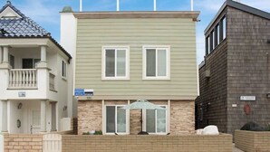 A two-story building with a light green facade, two windows with shutters, and a small patio area with a table and umbrella. - 114 31st A | Rare 1 bdr On Peninsula 3 Houses To Sand (Newport Beach)