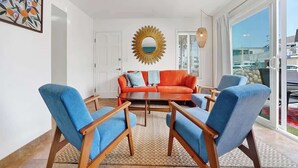 A living room with a sofa, two armchairs, a coffee table, a ceiling fan, and a large window with a view of the sea. - 117 31st St A | Stylish Renovation w/ Large Patio Steps To Sand (Newport Beach)