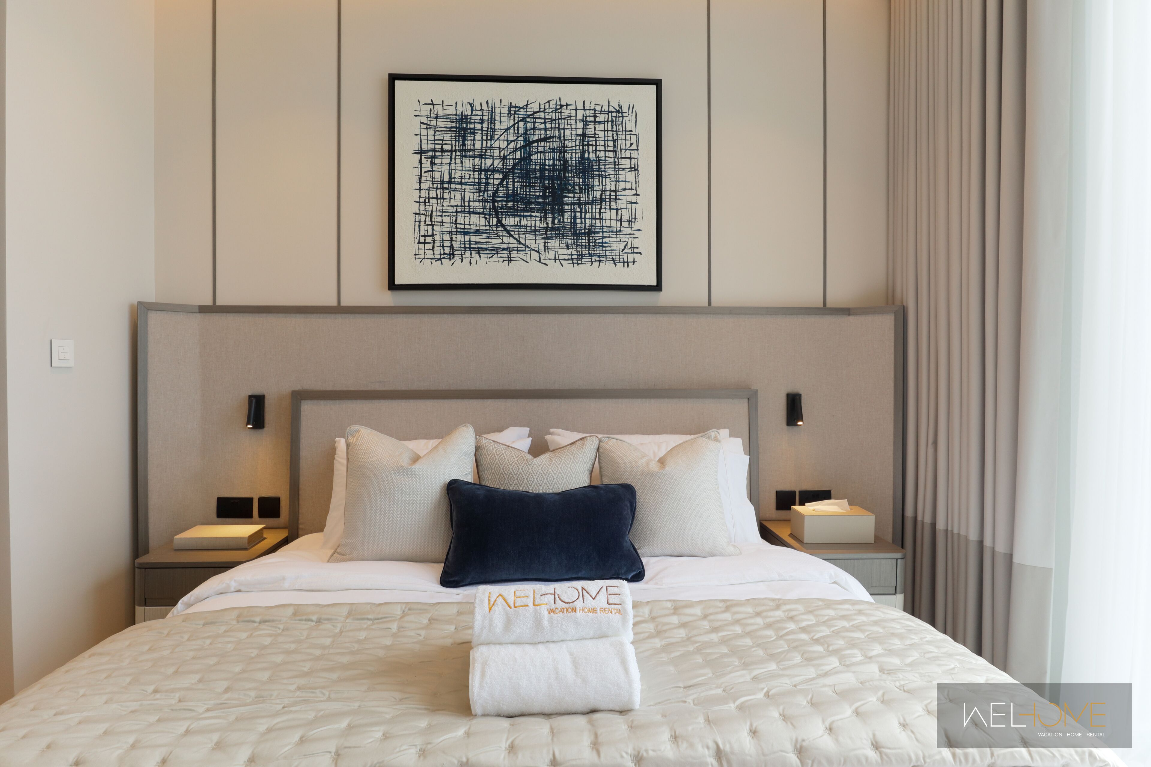 A neatly made bed with a white comforter and a folded towel with "WELCOME" text. There is a framed abstract artwork above the bed.