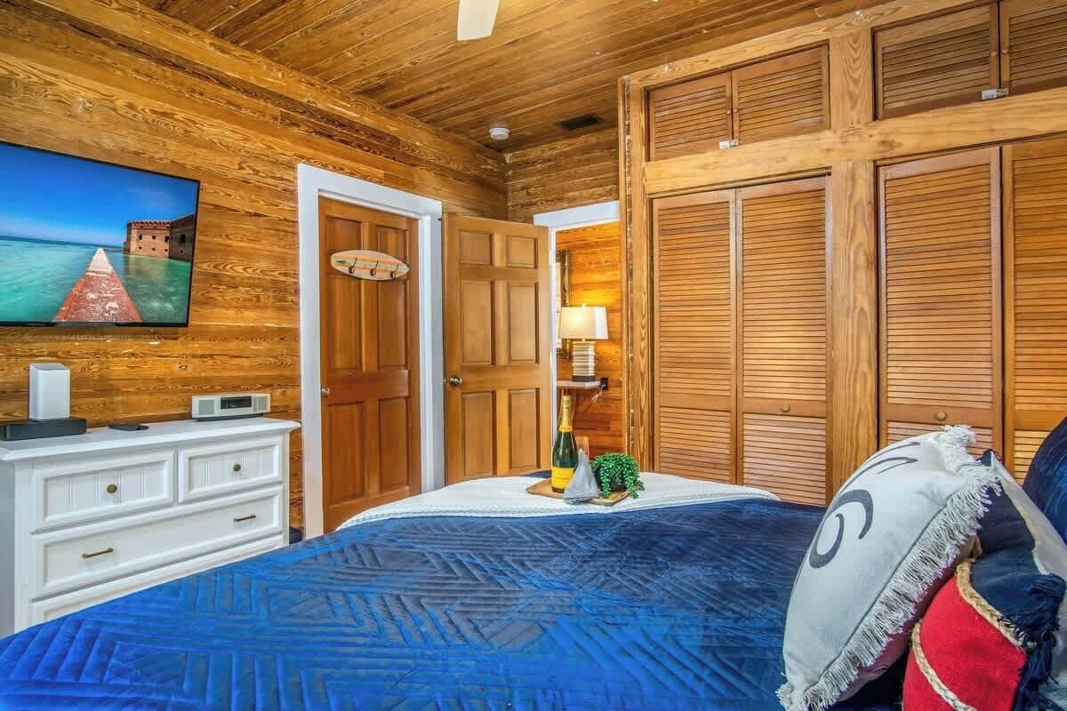 Una stanza con rivestimento in legno, un letto, una televisione, un comò e una porta che dà su un'altra stanza.