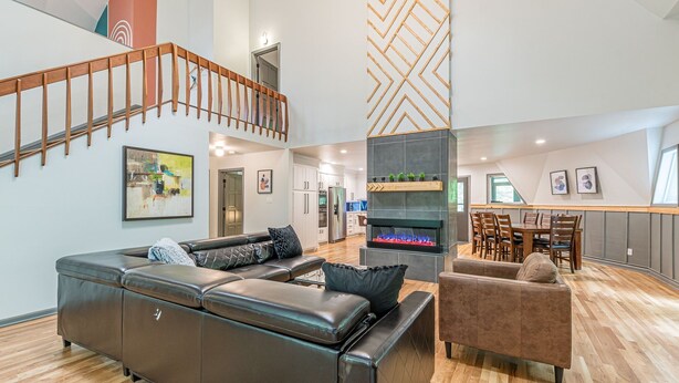 Un salón espacioso con un sofá de cuero marrón, un sillón marrón y una chimenea. Una escalera de madera conduce a un nivel superior.