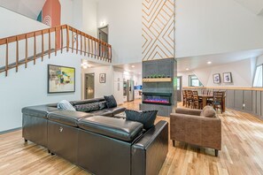 Un vaste espace de vie comprenant un canapé en cuir marron, un fauteuil marron et une cheminée. Un escalier en bois mène à l’étage supérieur.