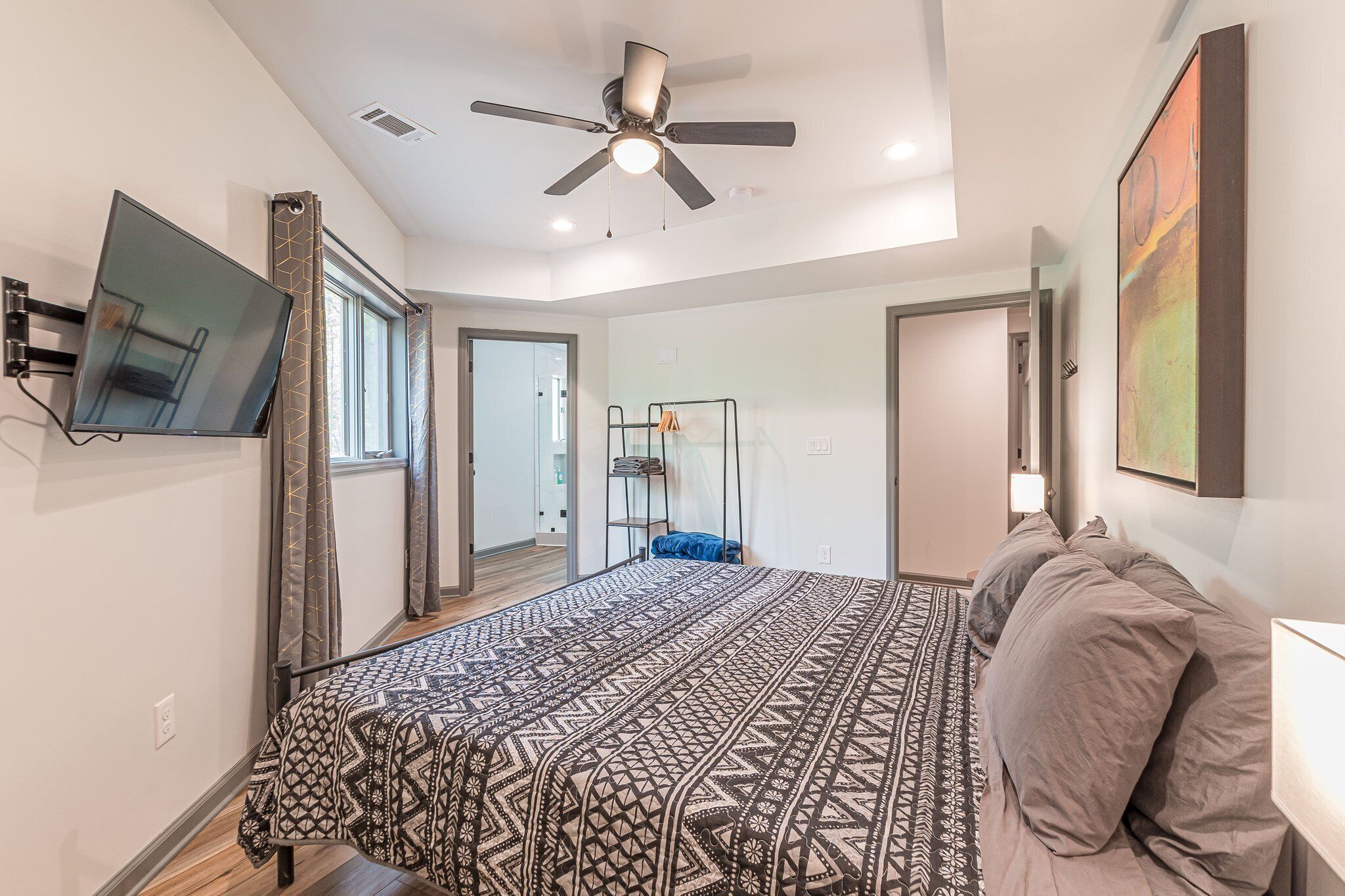 A bedroom with a bed, a ceiling fan, a TV mounted on the wall, and a door leading to another room.