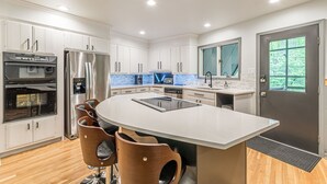 Una cocina moderna con armarios blancos, electrodomésticos de acero inoxidable, una isla grande con taburetes y una puerta oscura con ventana.