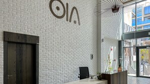 A reception area with a white brick wall, a reception desk, a dark wooden door, and a large window with a view of buildings. - WelHome - Prime 1BR Apartment at Oia Residence (Dubai)