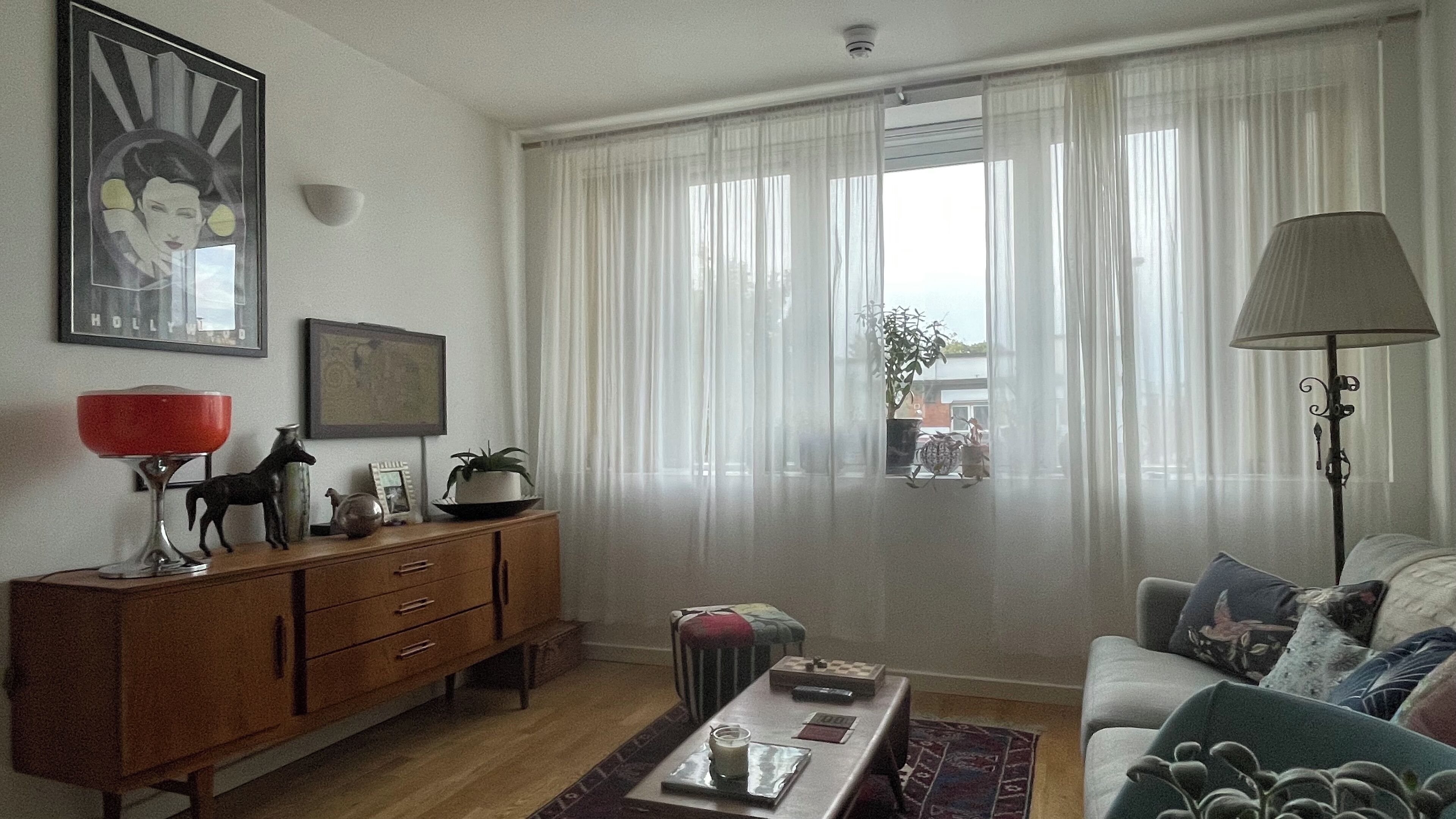 A living room with a wooden cabinet, a red lamp, a sofa with patterned cushions, and a window with sheer curtains.