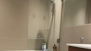 Moderni kylpyhuone, jossa on lasiseinällä varustettu suihku, valkoinen wc-istuin ja puinen allaskaappi.