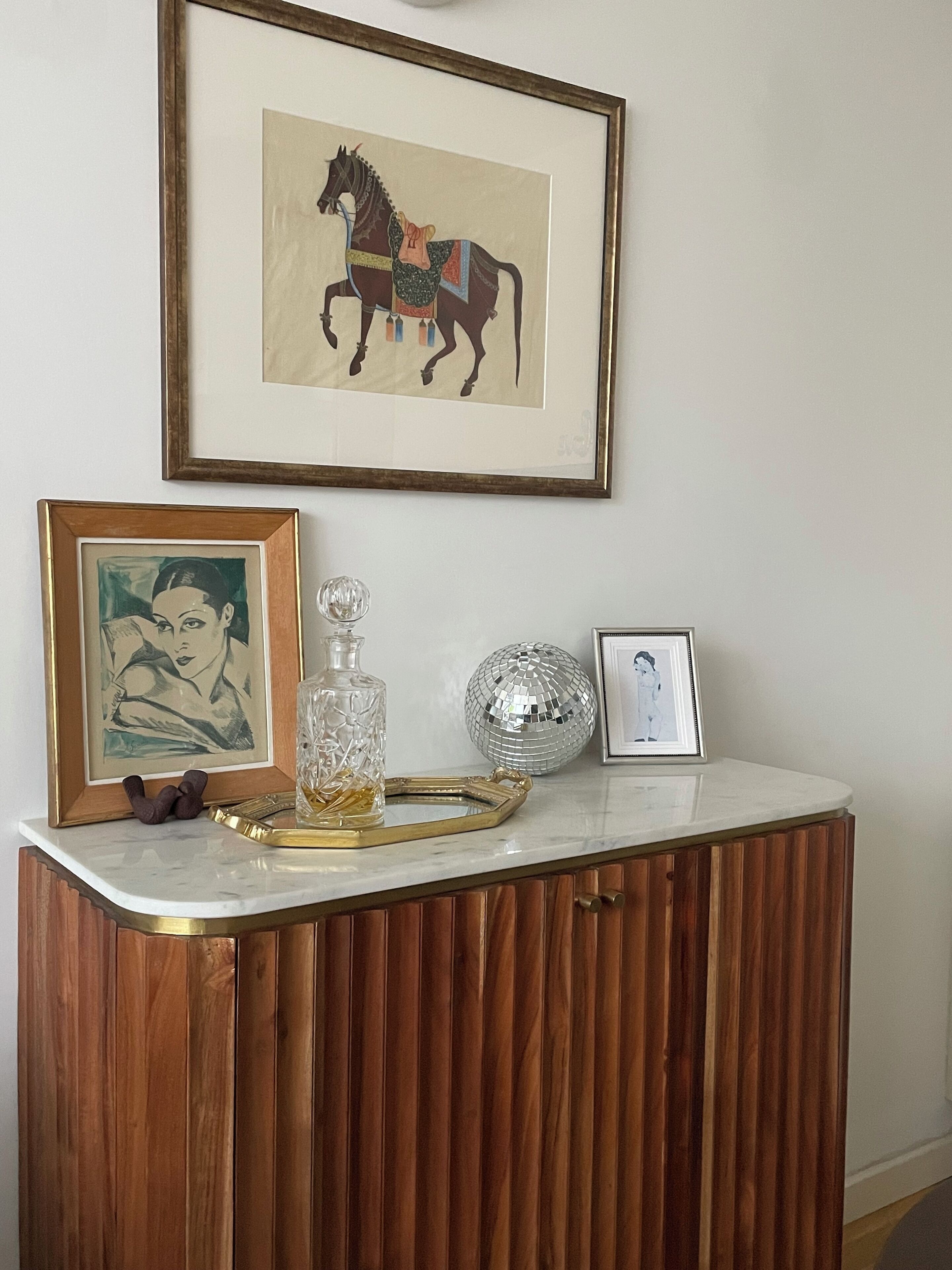Un meuble en bois surmonté d’un plateau en marbre, contenant des œuvres d’art encadrées, une carafe en cristal, une boule à facettes et un plateau.