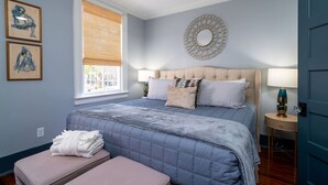 A bedroom with a large bed, a bedside table, a window with blinds, and wall art.
