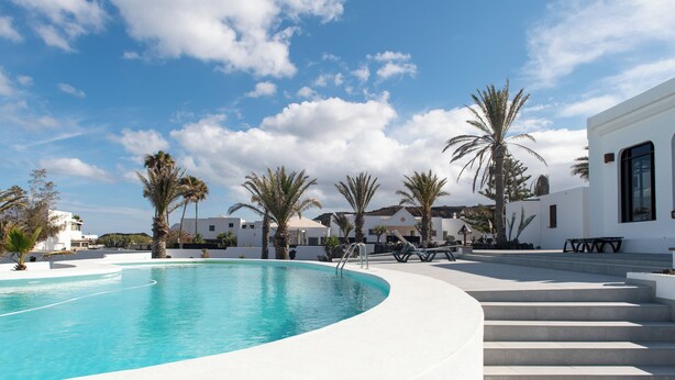 A pool area with a curved edge, palm trees, and a white building with steps leading up to a patio. - Villa Blanca 2, FKK - Lanzarote, Charco del Palo, Schönes Schwimmbad (Charco del Palo)