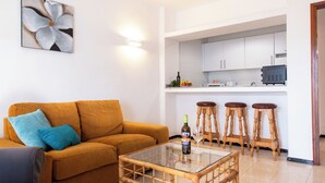 A living room with a sofa, a glass coffee table, and a kitchen area with a countertop and stools. - Villa Blanca 2, FKK - Lanzarote, Charco del Palo, Schönes Schwimmbad (Charco del Palo)