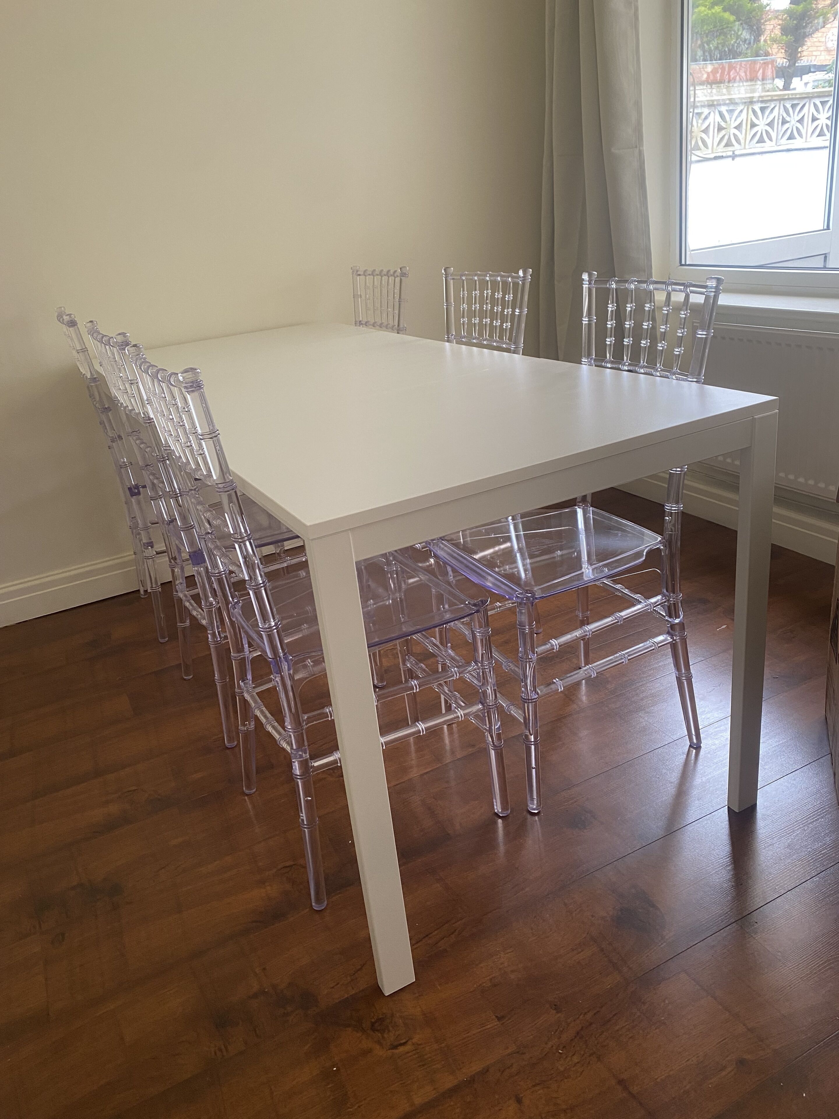 A white dining table with clear chairs in a room with wooden flooring and a window with a view of a fence.