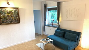 A modern living room with a blue sofa, a round ottoman, a small table with a vase, and a painting on the wall. - Modernes Zentrumsnahes Apartment (Oldenburg)