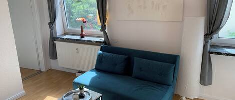 A modern living room with a teal sofa, a small side table, a radiator, and a window with curtains.