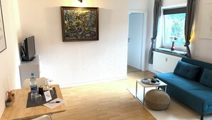 A modern living room with a wooden floor, a blue sofa, a dining table with chairs, a painting on the wall, and a window with curtains. - Modernes Zentrumsnahes Apartment (Oldenburg)