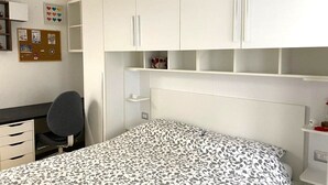 Un dormitorio con cama, escritorio y silla. Hay armarios blancos sobre la cama y un tablón de anuncios en la pared.