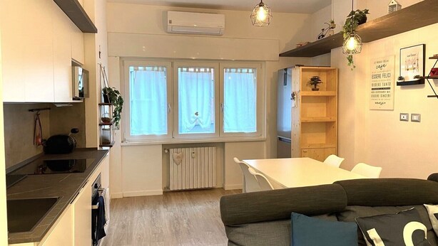 Una cocina moderna con horno empotrado, un comedor con mesa y sillas blancas, y una sala de estar con sofá y cojines.