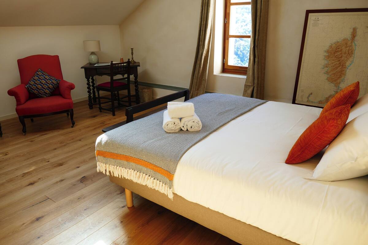 A bedroom with a large bed, a red armchair, a small table with a lamp, and a map on the wall.
