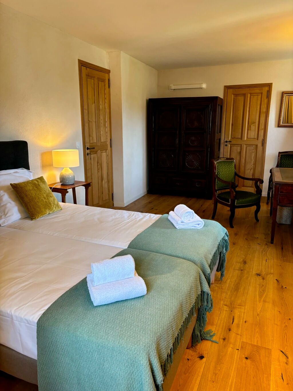 A hotel room with a large bed, two bedside tables with lamps, a wooden wardrobe, a green armchair, and a desk with a chair.
