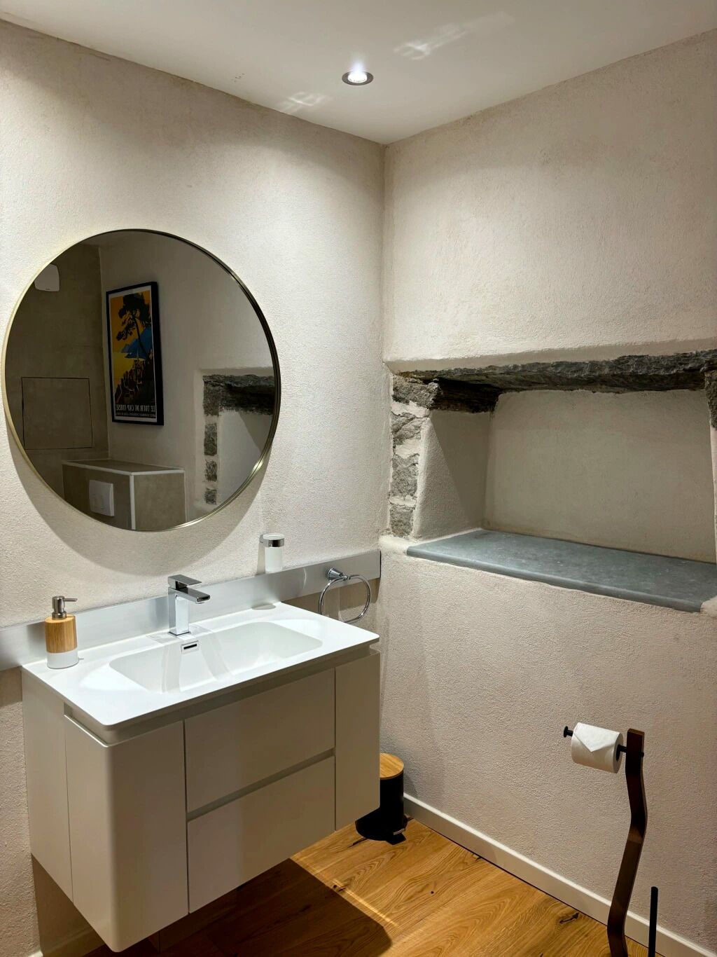 Ein modernes Badezimmer mit weißem Waschtisch, einem runden Spiegel, Holzboden und einer Wand mit Steinverkleidung.