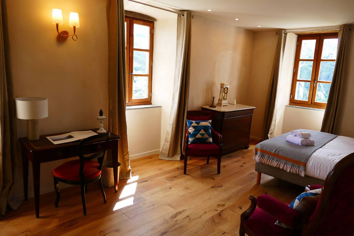 A bedroom with a wooden floor, a desk with a lamp, a chair, a bed with a blanket, and two windows with curtains.