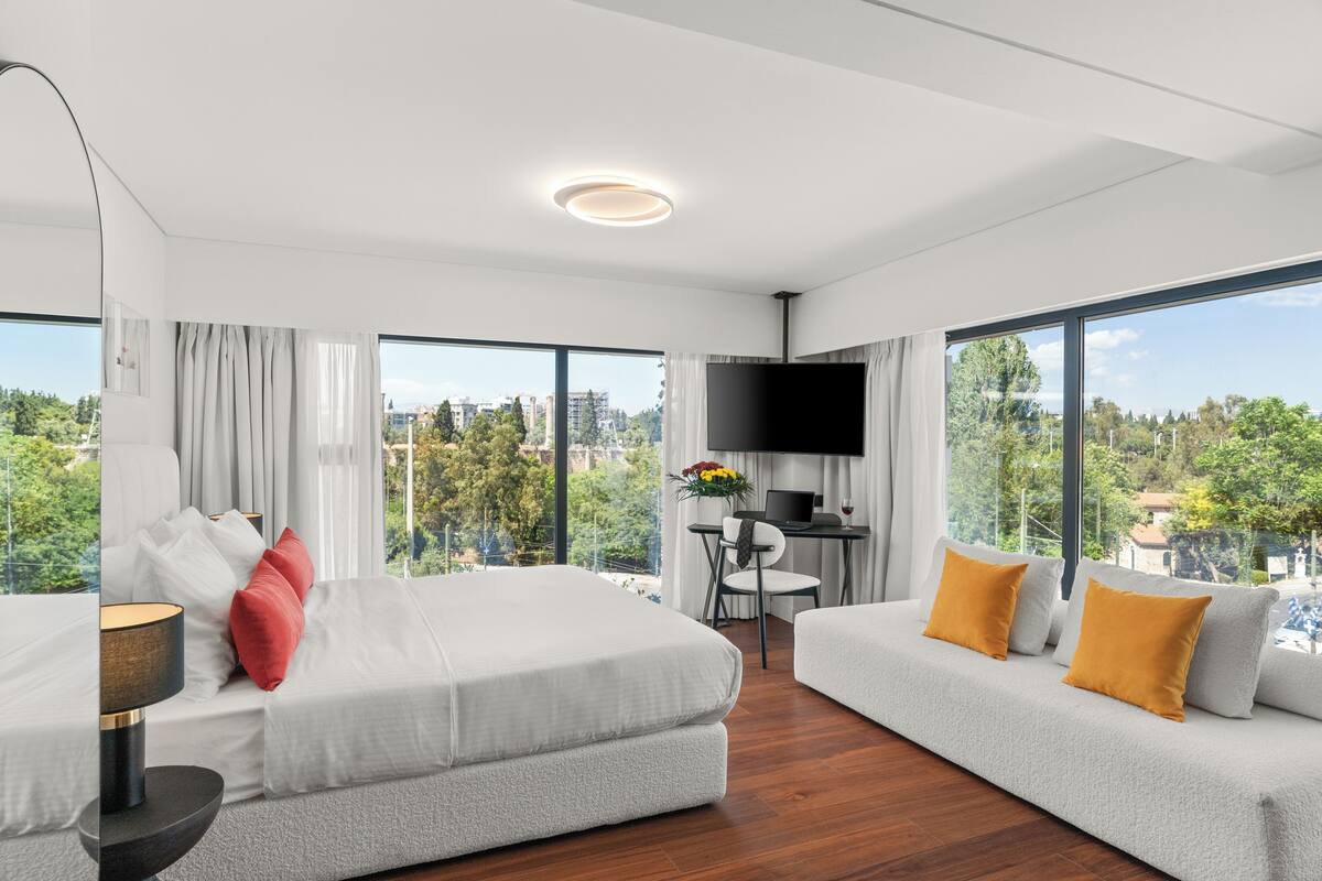 A modern hotel room with a large bed, a sofa, a desk with a computer, and a TV.