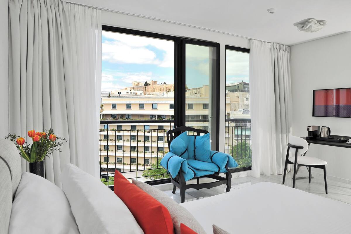 A modern hotel room with a large balcony, a sofa, a blue armchair, and a dining area with a TV.