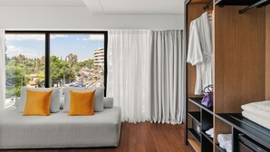 A modern hotel room with a large window, a grey sofa with orange cushions, a wooden wardrobe, and a view of a cityscape. - The Athenaeum Luxury Hotel (Athens)