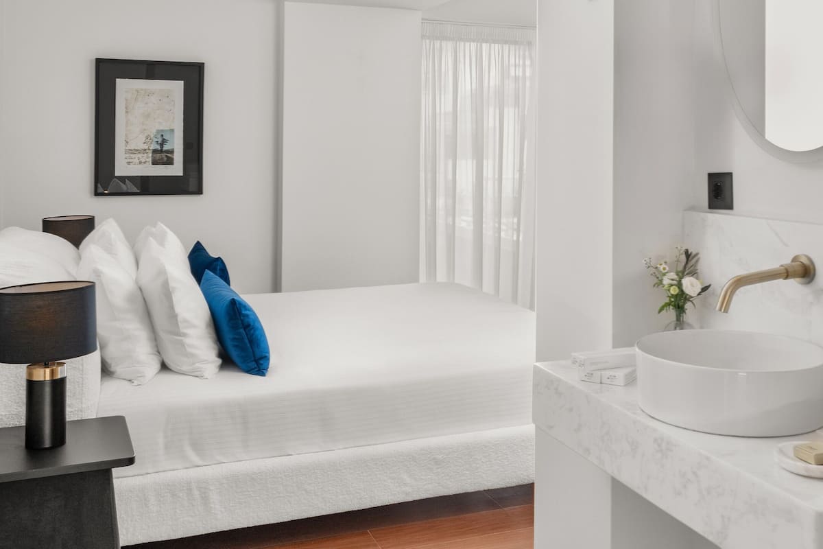 A modern bedroom with a white bed, a bedside table with a lamp, a framed picture on the wall, and a bathroom with a round sink and mirror.