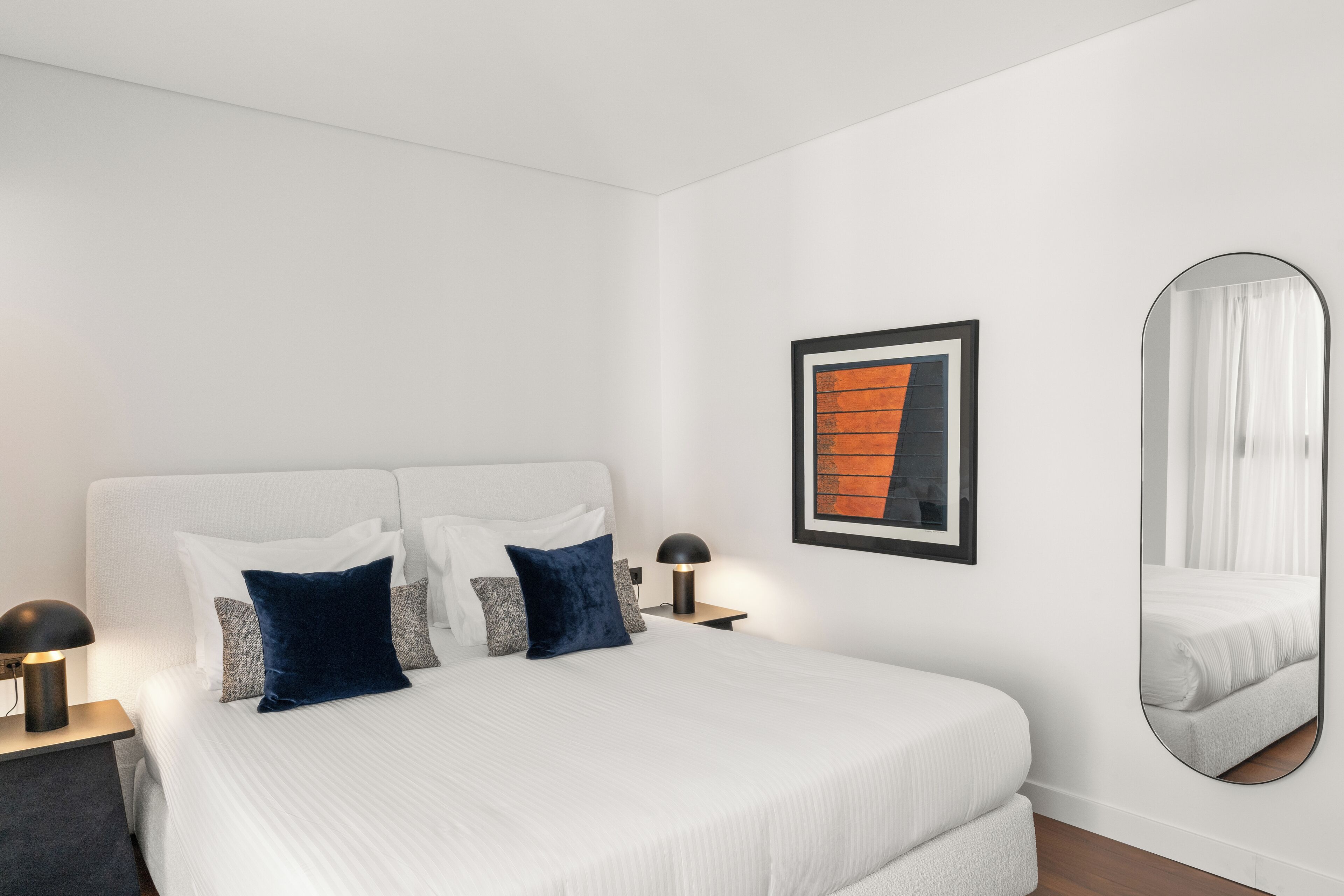 Um quarto bem arrumado, com uma cama grande, uma mesa de cabeceira com abajur, um quadro na parede e um espelho.