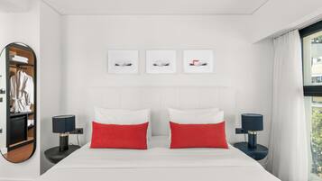 Quarto de hotel moderno com uma cama grande, duas almofadas vermelhas, uma cabeceira branca e três quadros na parede.