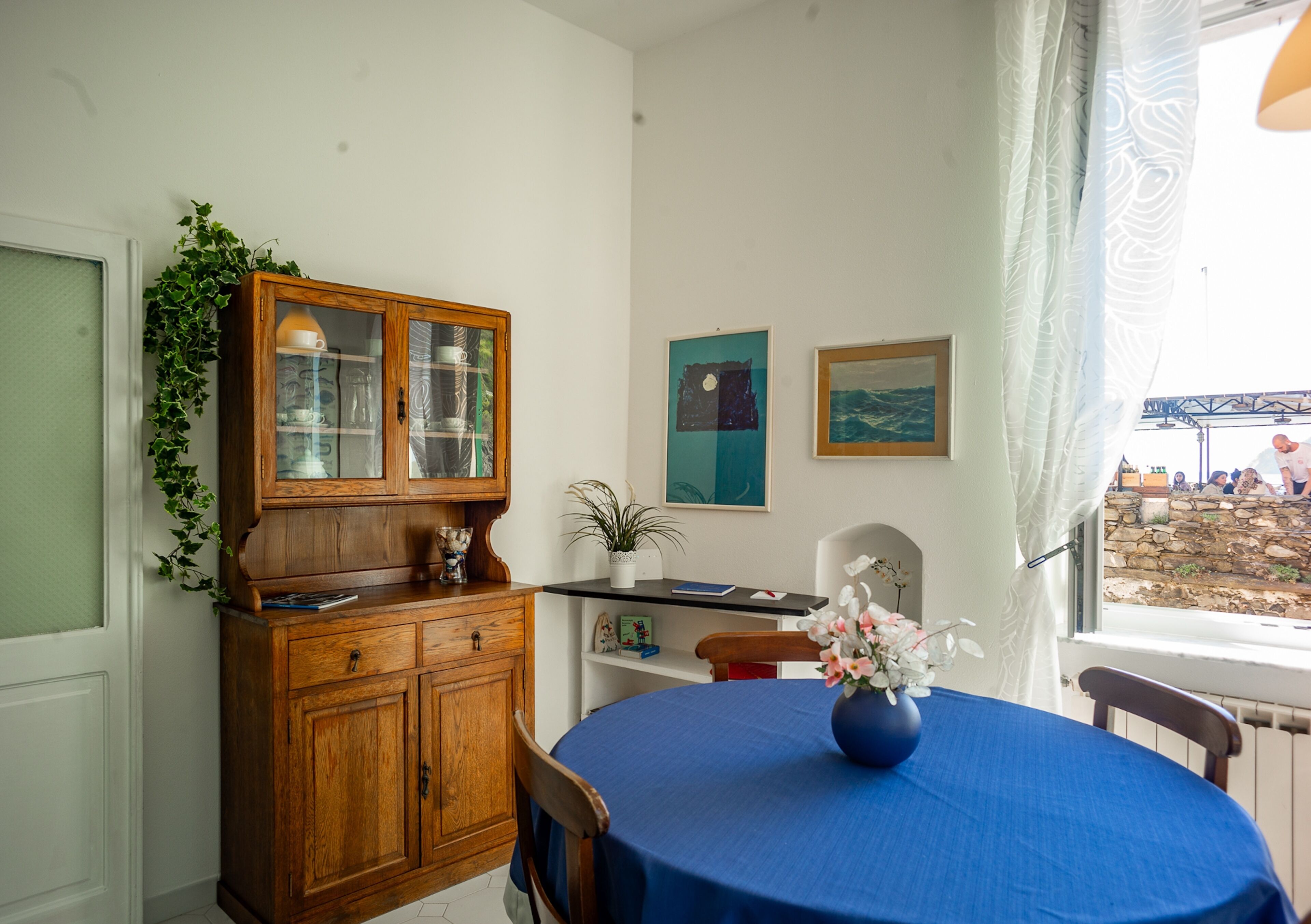 Een eethoek met een ronde tafel bedekt met een blauwe tafelkleed, een houten kast met glazen deuren en een potplant.