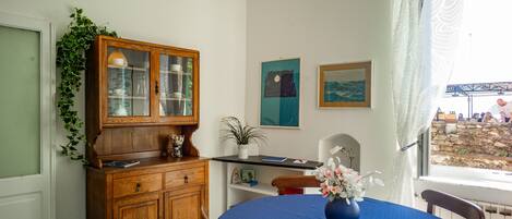 A dining area with a round table covered in blue tablecloth, a wooden cabinet with glass doors, and a potted plant.