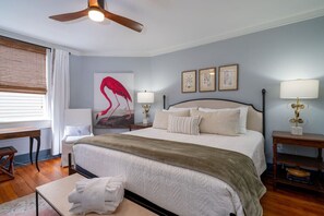 A bedroom with a bed, bedside tables, a desk, a chair, a ceiling fan, and artwork on the walls.