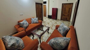 Un salon avec des canapés orange, des coussins à motifs, une table basse en verre, un ventilateur de plafond et un réfrigérateur rouge.
